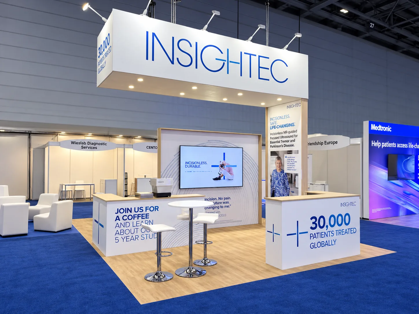 Stand d’exposition sur mesure en îlot réalisé par Mydisplay pour Insightec, avec bandeau suspendu, plancher en bois, comptoirs d’accueil et bar à café